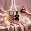 Perfume bottle+box on a vanity table draped in soft nude silk, tuberose blossoms scattered, soft pink light, mirror reflections. Woman (hijab optional) gently sprays