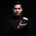 Malay man in black shirt presenting Suqraat perfume, sharp contrast lighting, cinematic men's product style