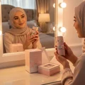 Malay hijabi woman holding perfume in front of mirror with warm light and soft mist – elegant women fragrance
